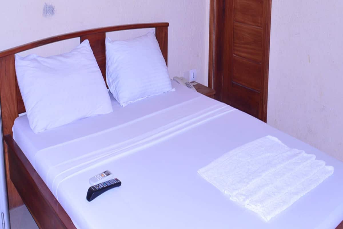 Una cama bien hecha con sábanas blancas, un control remoto y una toalla blanca doblada sobre un cabecero de madera en una habitación con puerta de madera y paredes de tonos claros.