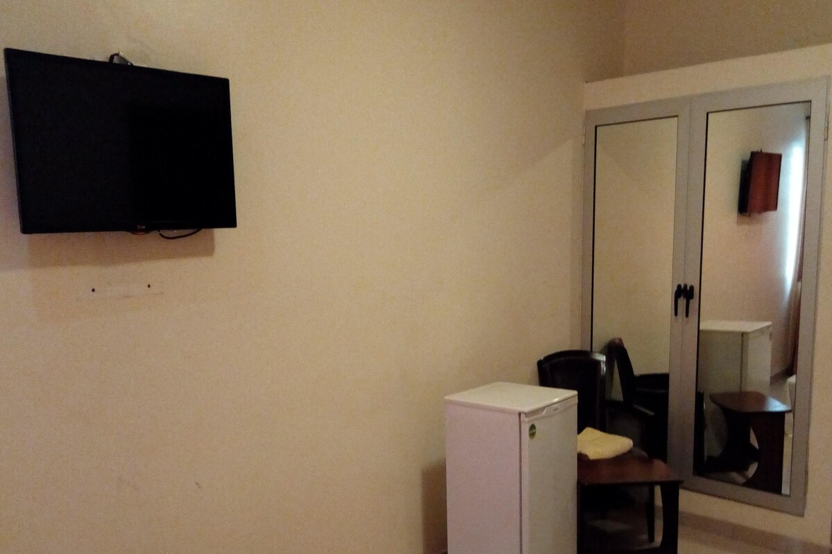 A hotel room with a bed, a small white refrigerator, a television mounted on the wall, and a mirror in the door.