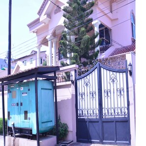 Un edificio grande con entrada protegida por una reja, un cuadro eléctrico azul y un árbol en primer plano.