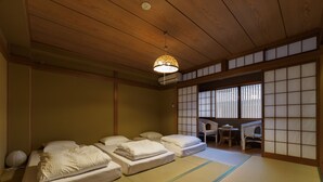 Une pièce de style japonais traditionnel, avec un sol recouvert de tatamis, un plafond bas en bois et des paravents coulissants en papier de riz (shoji). On y trouve deux tatamis avec des coussins, ainsi qu’une petite table et une chaise.