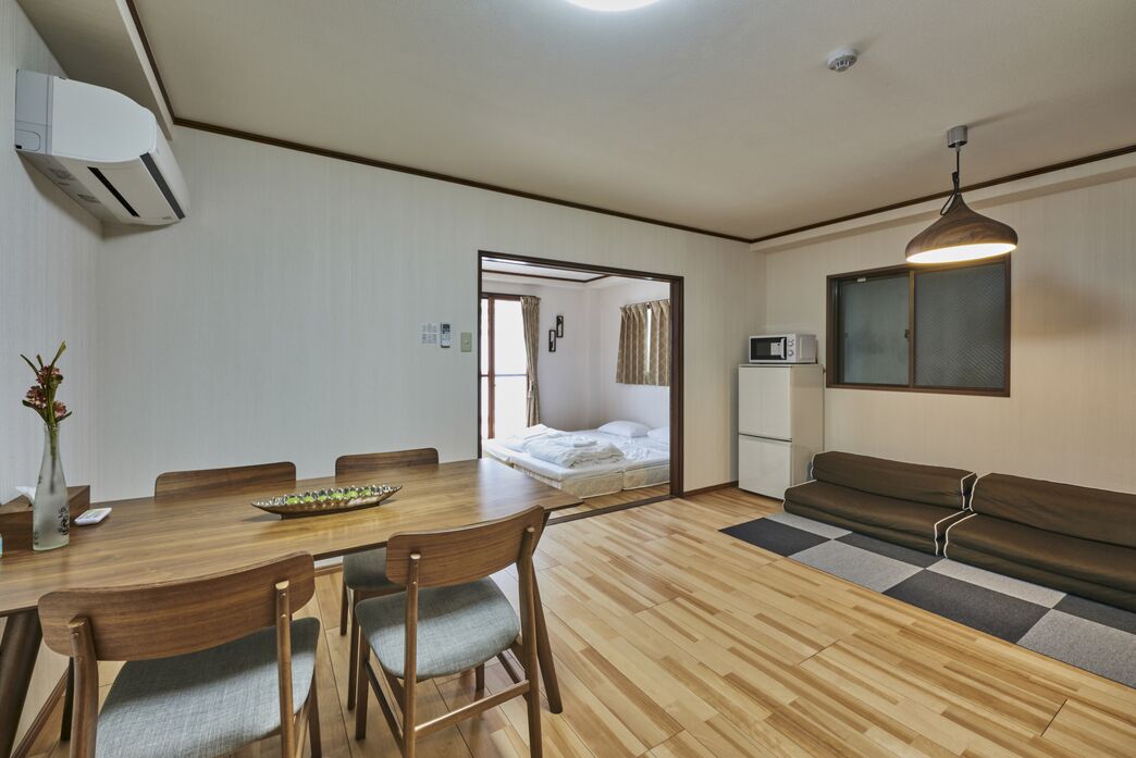 A compact room with a wooden dining table, chairs, a sofa, a bed, and a kitchenette.