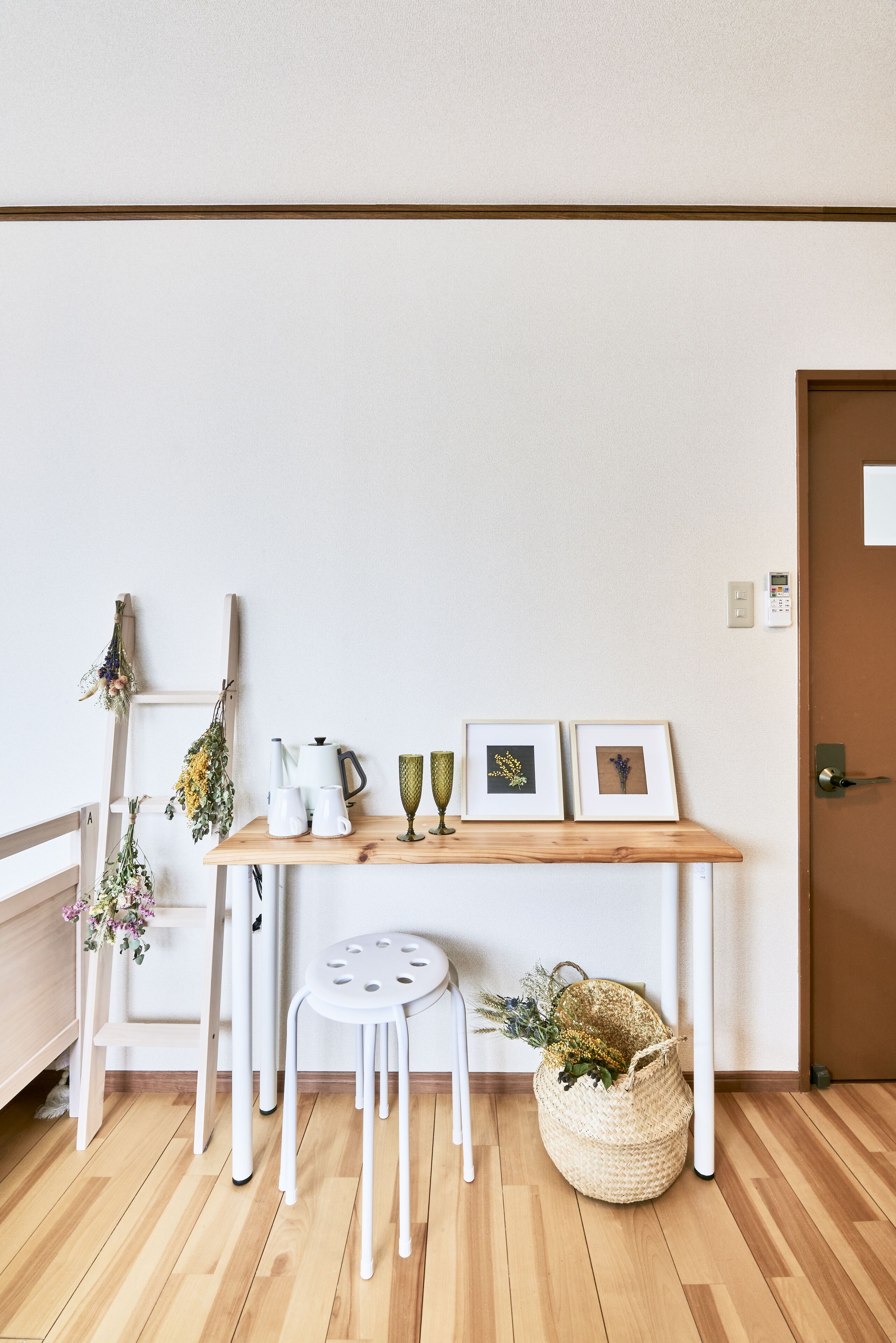 A room with a wooden floor, a white table with a white chair, a small white stool, a woven basket, and a door with a handle.