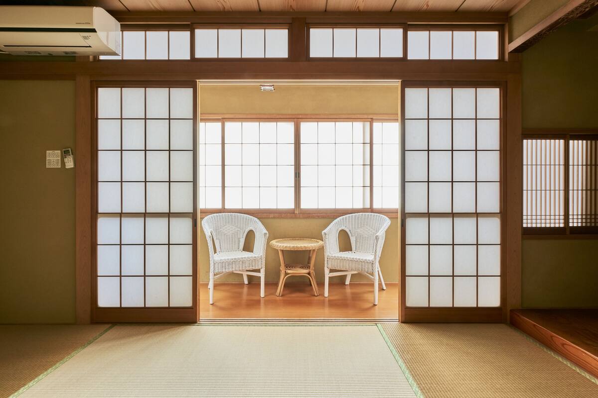 Une pièce de style japonais traditionnel, avec un sol recouvert de tatamis, une porte coulissante en papier de riz (shoji) et deux chaises en osier blanc disposées autour d’une petite table.
