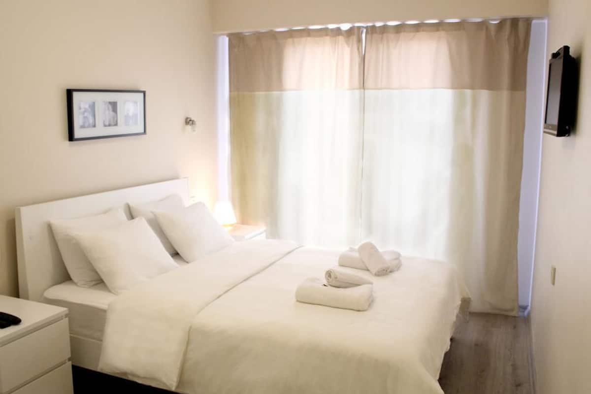 A hotel room with a bed, white bedding, a bedside table, a framed picture on the wall, and a window with curtains.