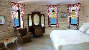 A bedroom with a large bed, a wooden dresser, a chair, and a small table. There are two windows with floral curtains and a view of the sea. - Nemrut Bay Otel (Foça)