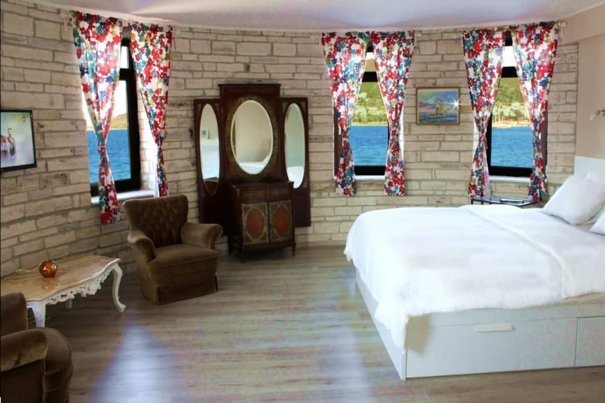 A bedroom with a large bed, a wooden dresser, a chair, and a small table. There are two windows with floral curtains and a view of the sea.