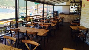 An indoor dining area with wooden tables and chairs, large windows offering a view of the beach and sea, and a bar area with bottles on shelves. - Nemrut Bay Otel (Foça)