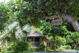 A bungalow with a thatched roof, surrounded by lush greenery and a sign indicating its name.