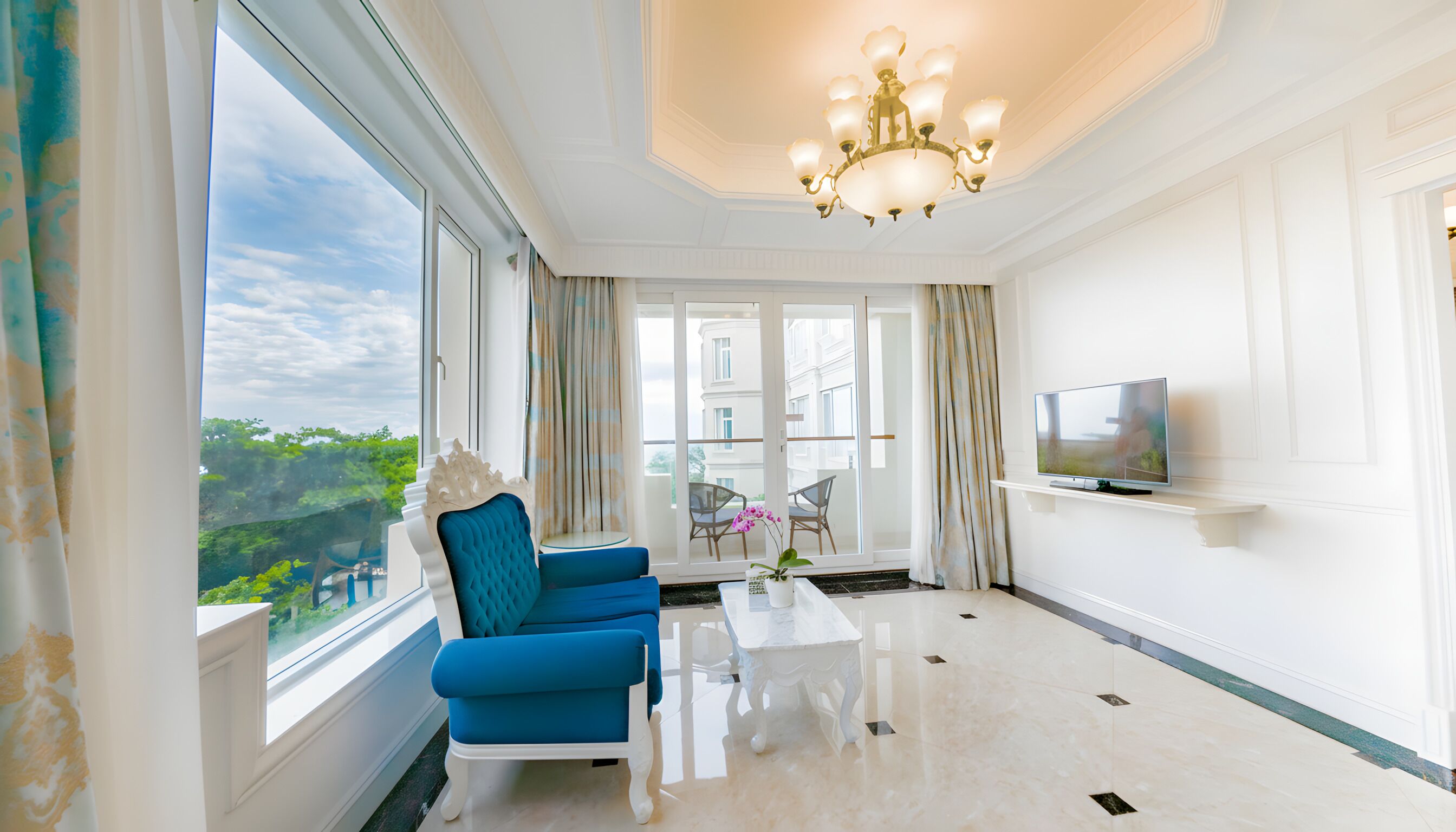 A spacious room with a large window, a blue sofa, a white coffee table, and a chandelier.