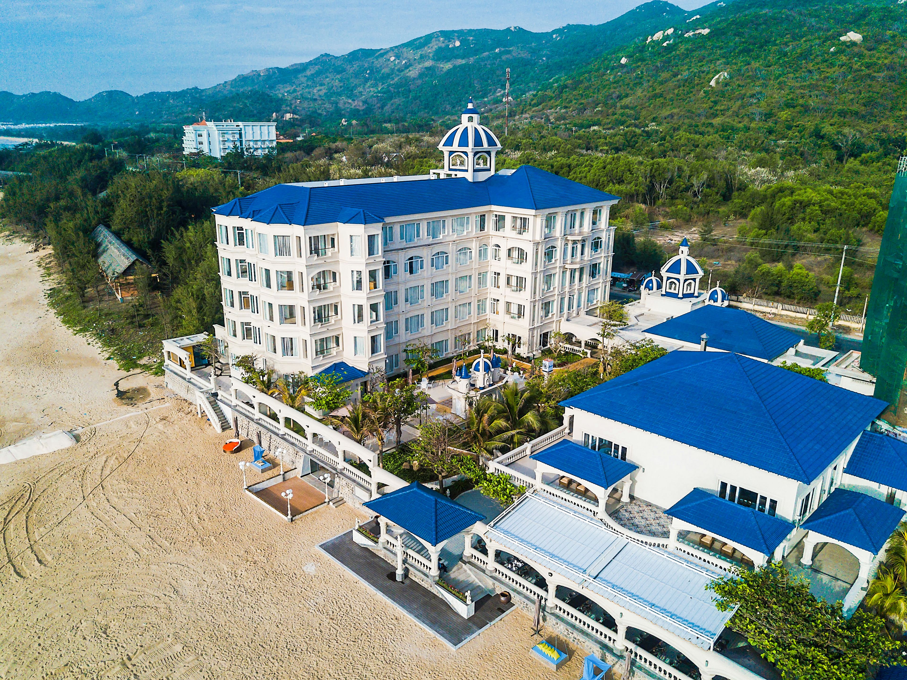 A large, multi-story hotel with a prominent blue dome and white exterior, situated on a beach with a mountainous backdrop.