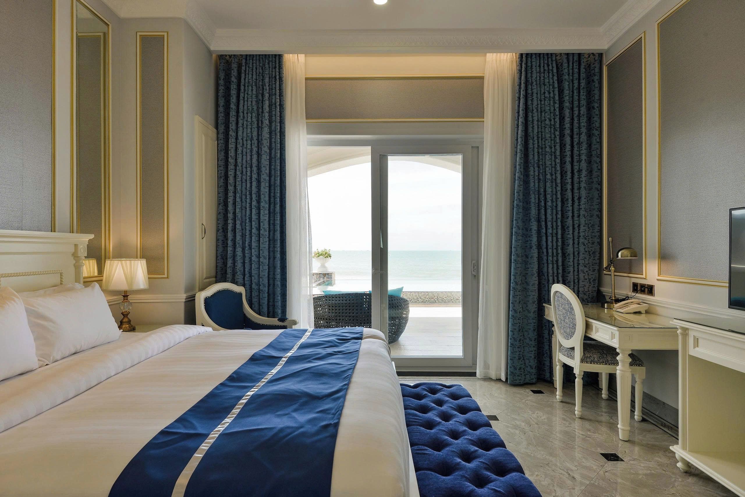 A hotel room with a large bed, a desk with a chair, a television, and a view of the sea.