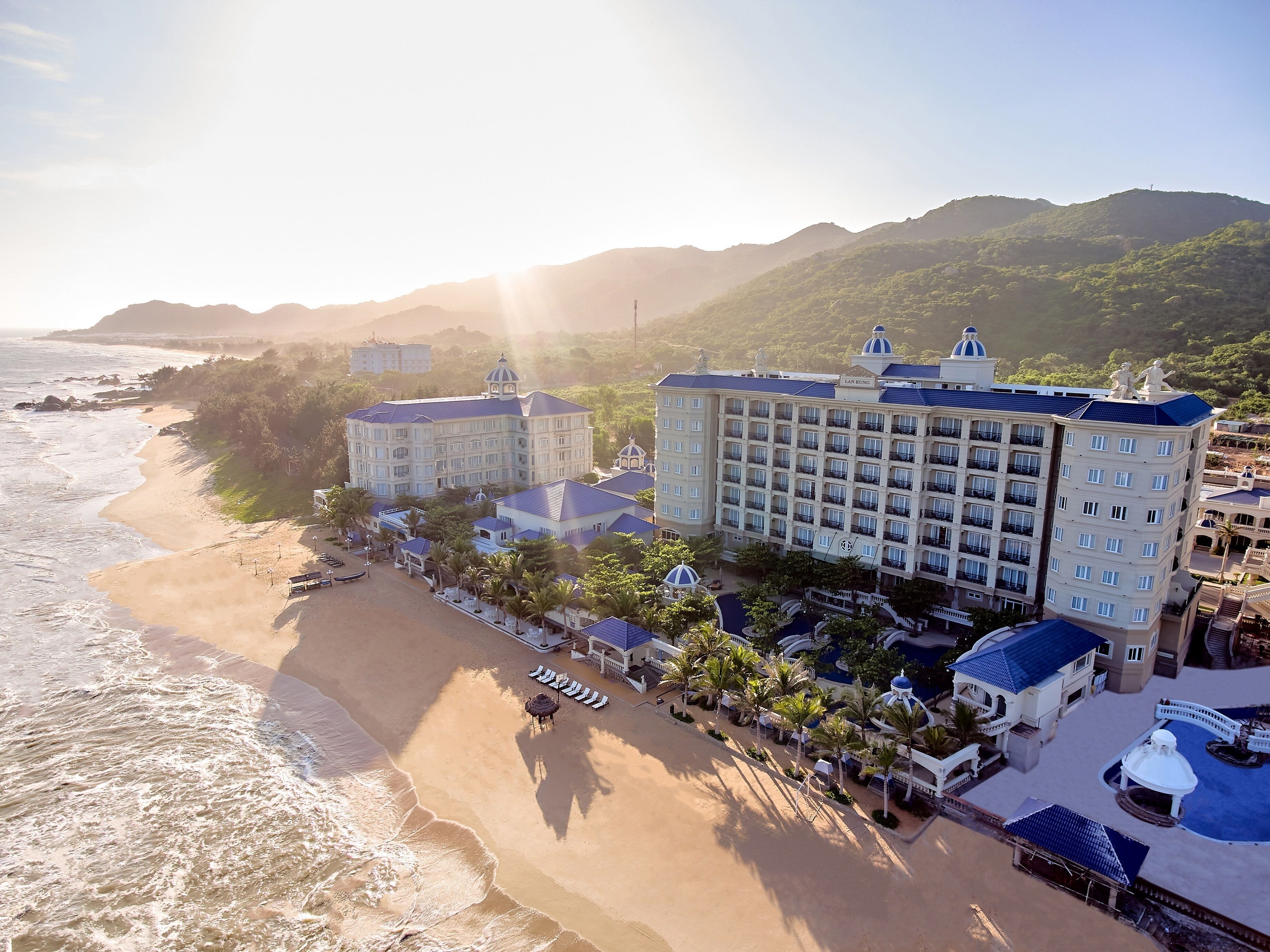 Un hotel frente a la playa con varias construcciones, una playa y montañas al fondo.