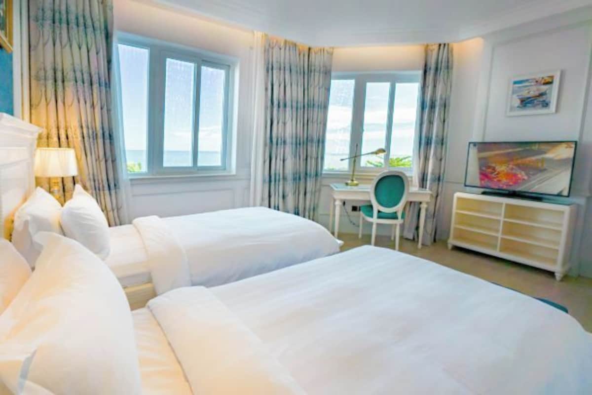 A hotel room with two beds, a desk, a chair, a TV, and a window with curtains.