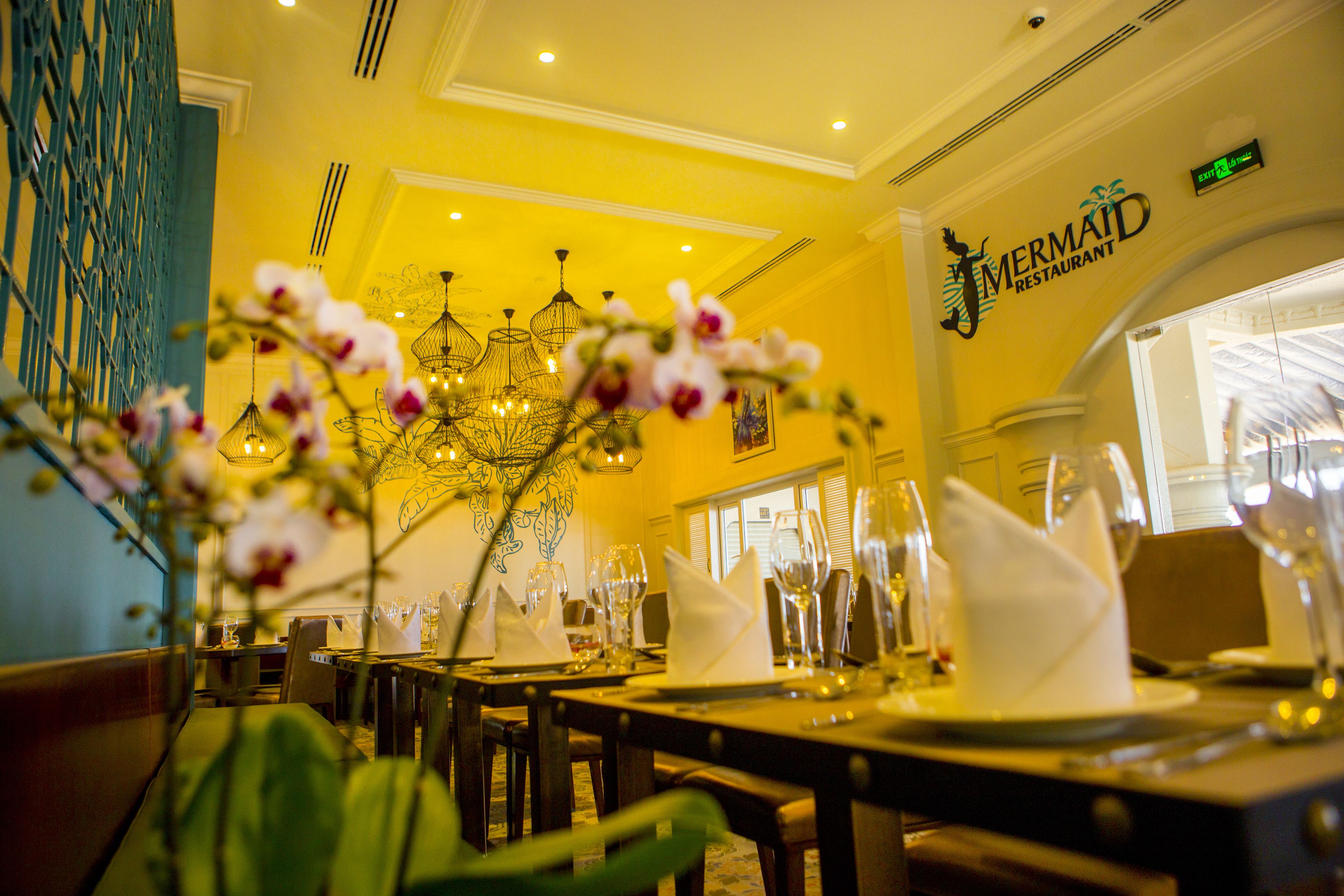 A restaurant interior with yellow walls, chandeliers, and neatly set dining tables.