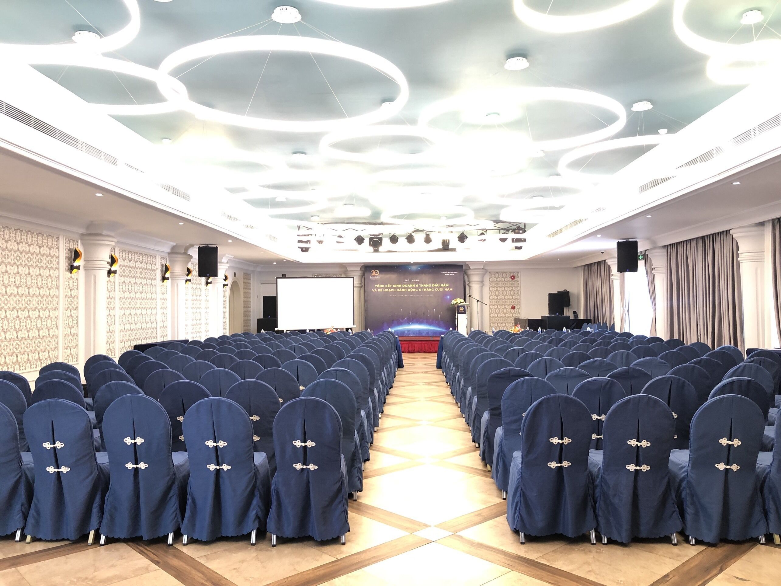 A conference room with rows of blue chairs, a stage with a podium, and a modern ceiling design.
