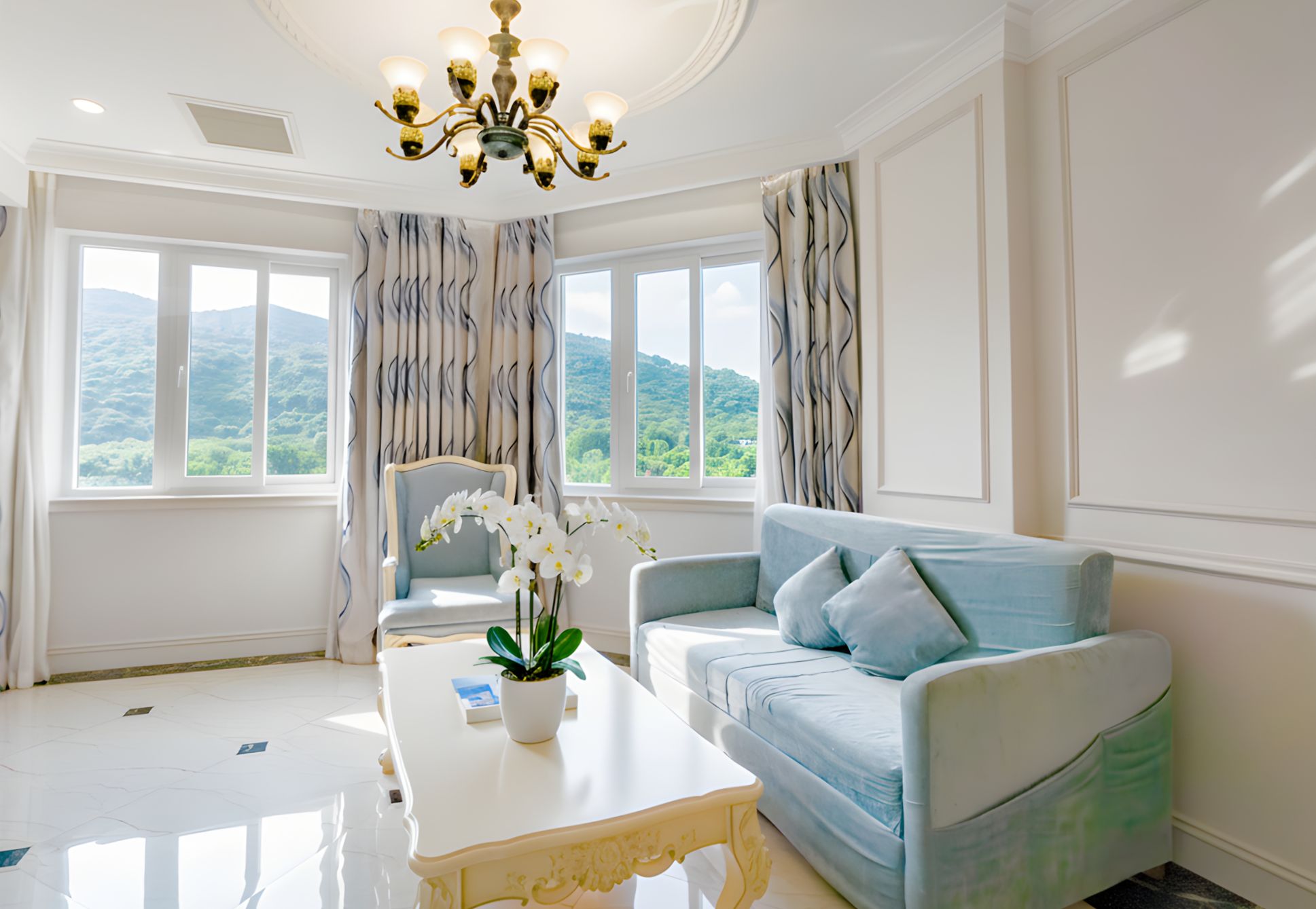 A room with a sofa, a coffee table, and a chair. There are windows with curtains, a chandelier, and a view of greenery outside.