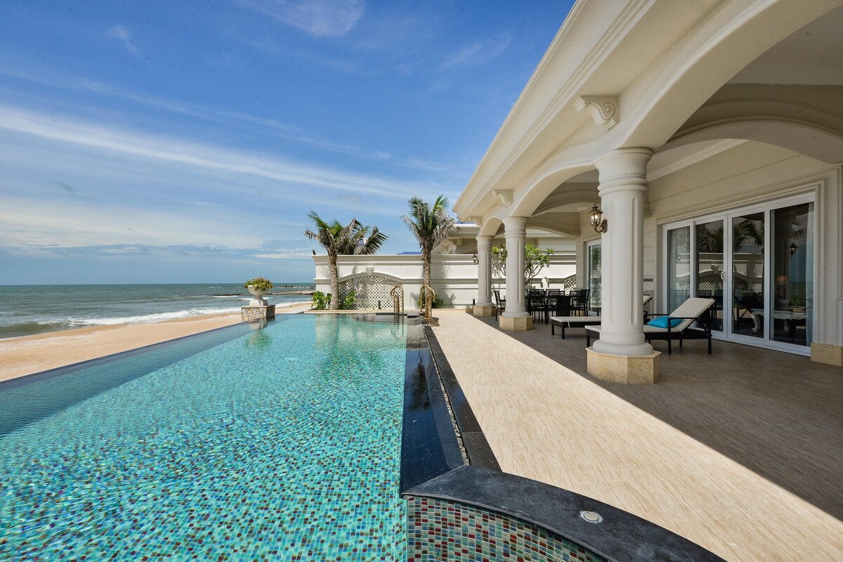A poolside area with a view of the ocean, a covered patio with seating, and a beach in the background.