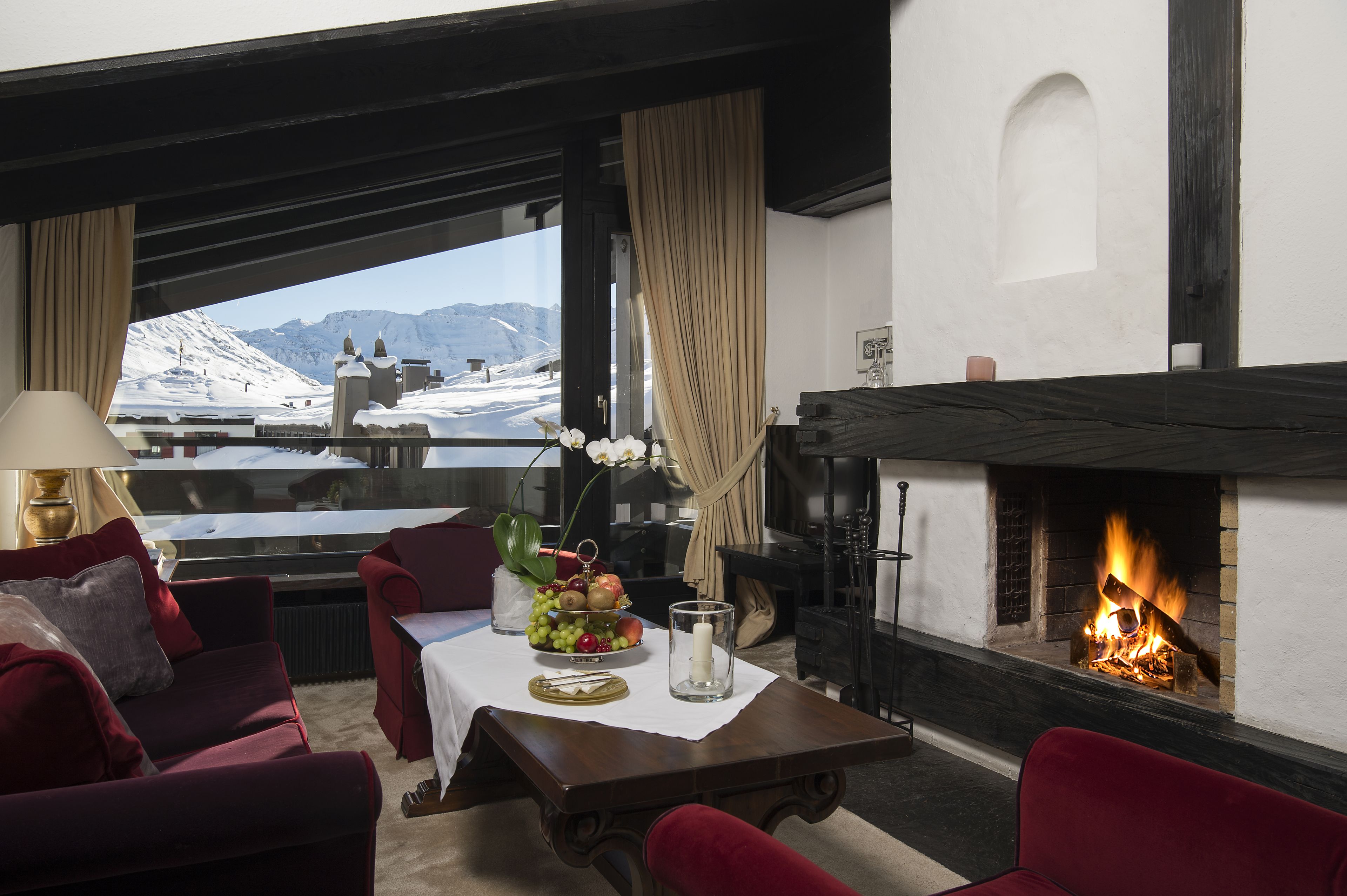 Una habitación acogedora con chimenea, una mesa con frutas y velas, y vistas a montañas nevadas.