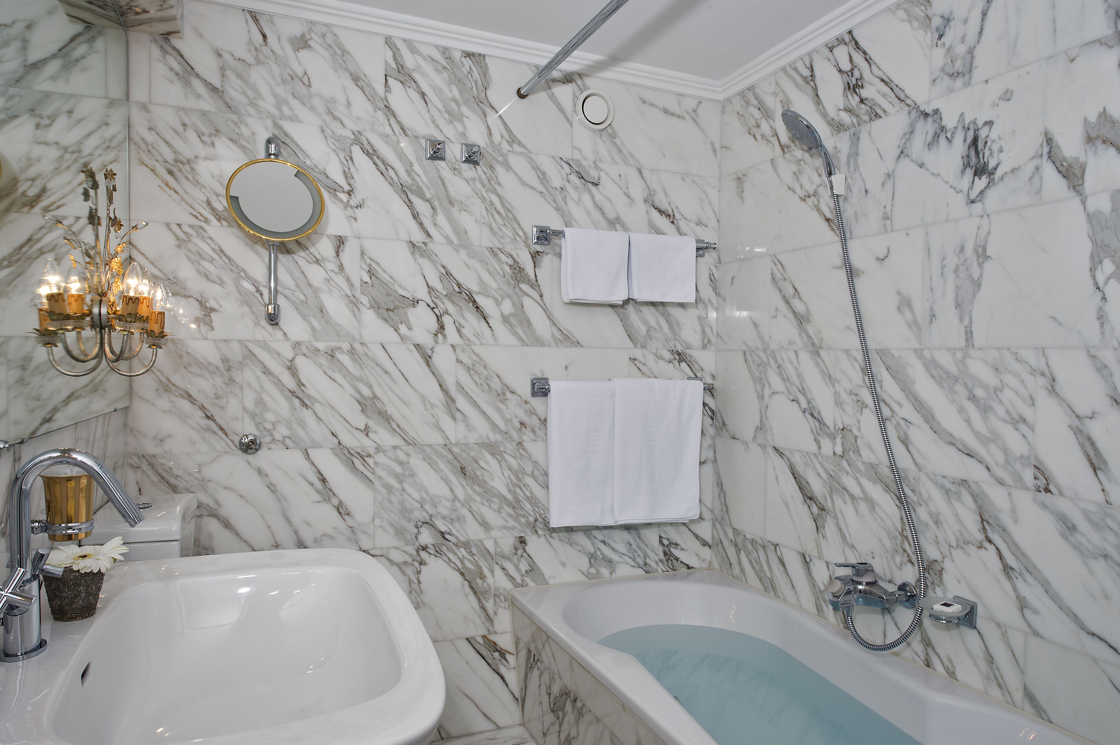A bathroom with marble walls, a bathtub, a sink, a mirror, and a chandelier.