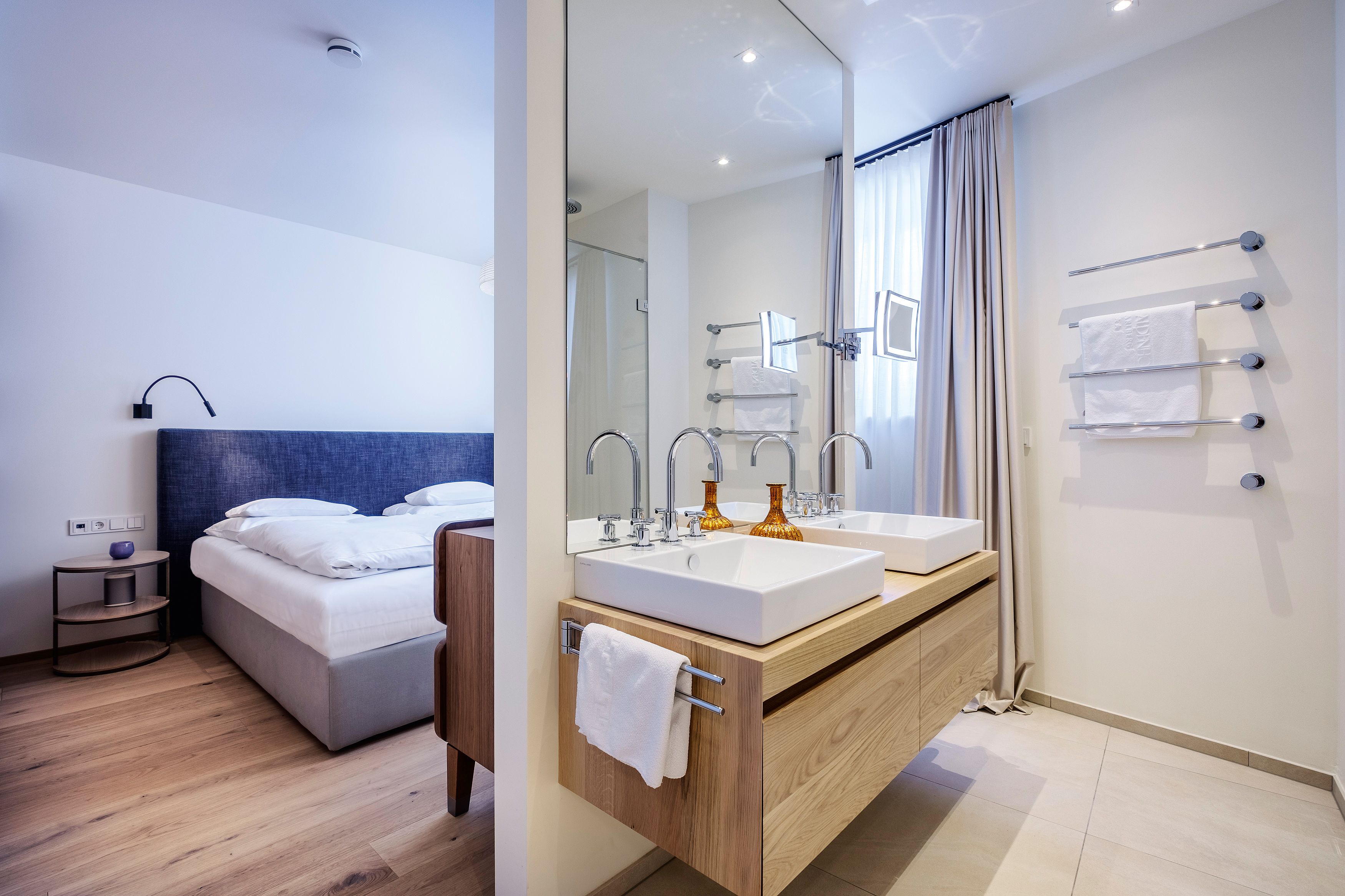 Une chambre d’hôtel moderne comprenant un lit, une table de chevet en bois, une salle de bain avec un double lavabo et un grand miroir.