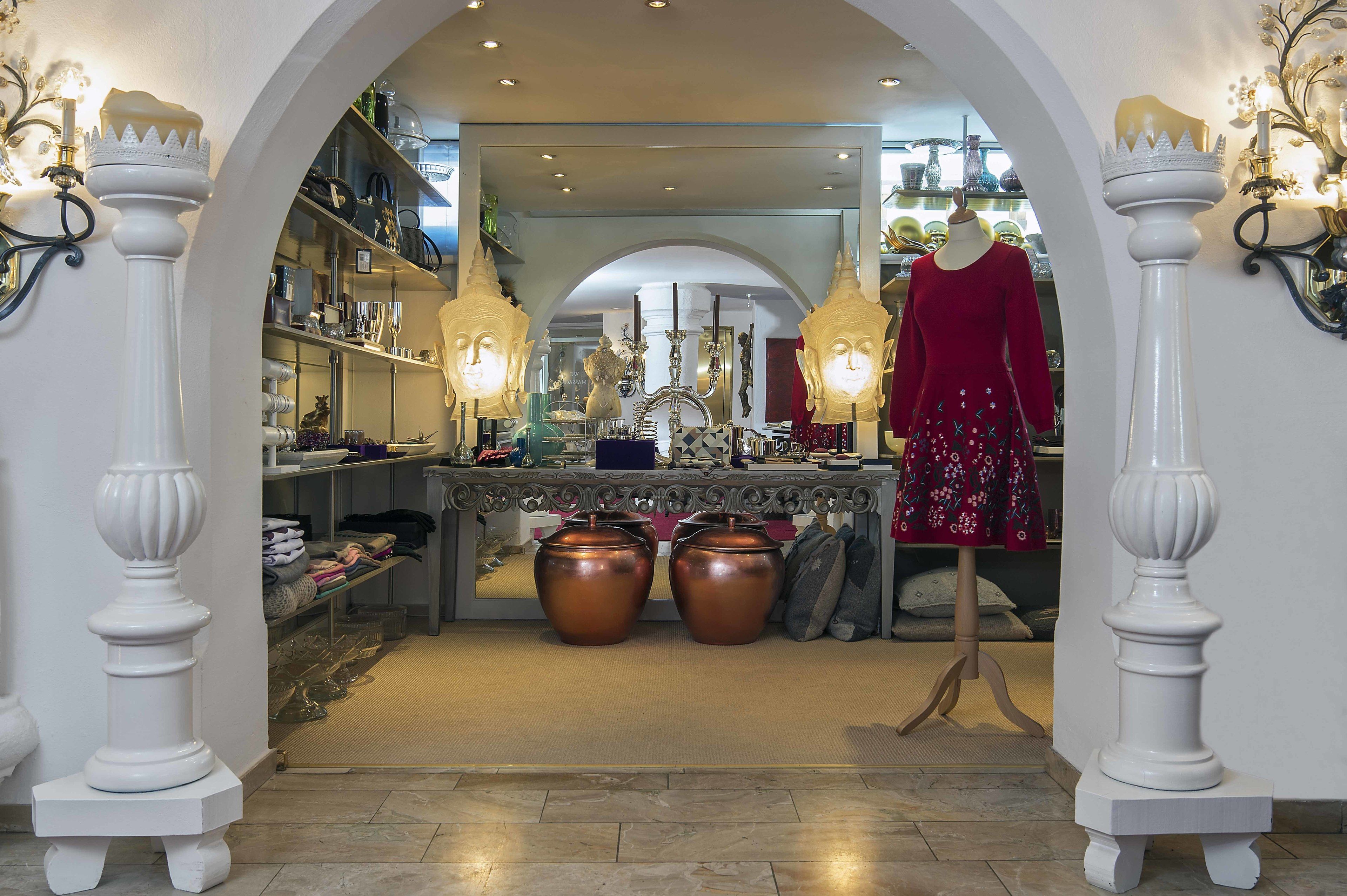 An interior with an arched doorway, shelves with various items, a mannequin in a red dress, and two large urns.