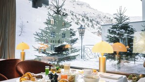 Una mesa de comedor con alimentos para el desayuno y vistas a montañas nevadas a través de una ventana.