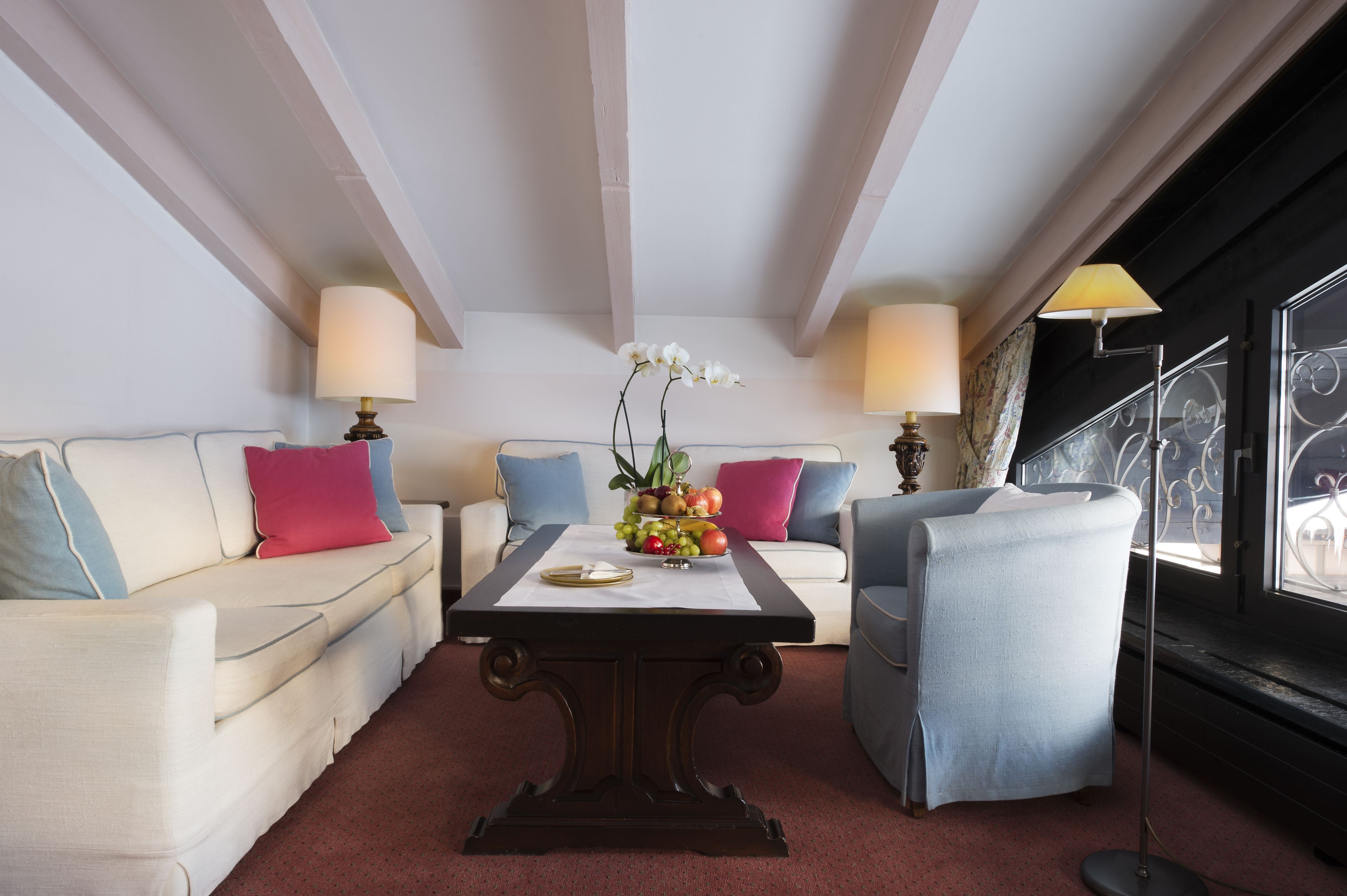 A room with a white sofa, a blue armchair, a wooden coffee table with a fruit centerpiece, and two lamps.