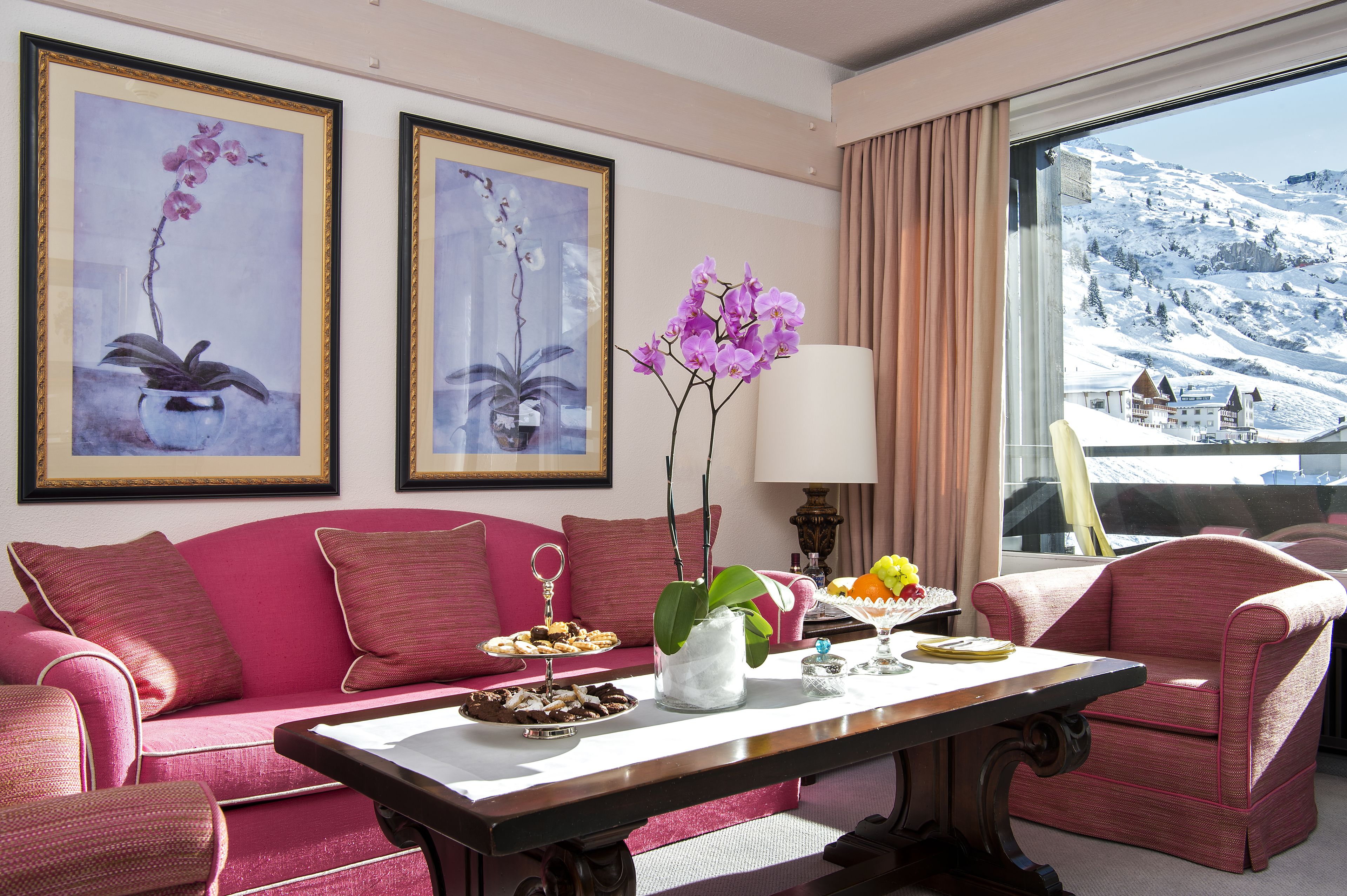A room with a pink sofa, a dining table with a vase of purple flowers, and framed artwork on the wall.