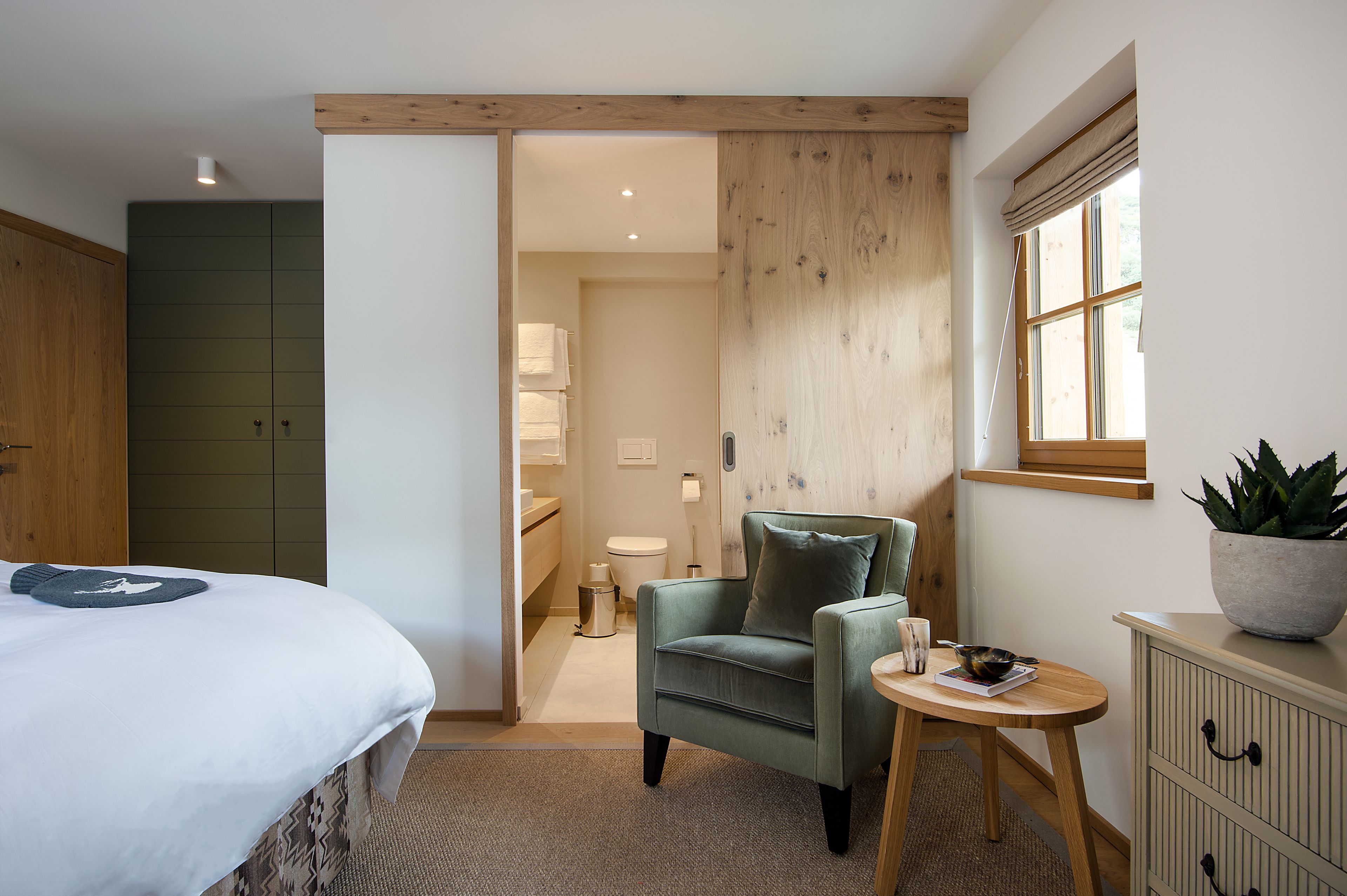 A bedroom with a bed, a chair, a small table, and a potted plant.