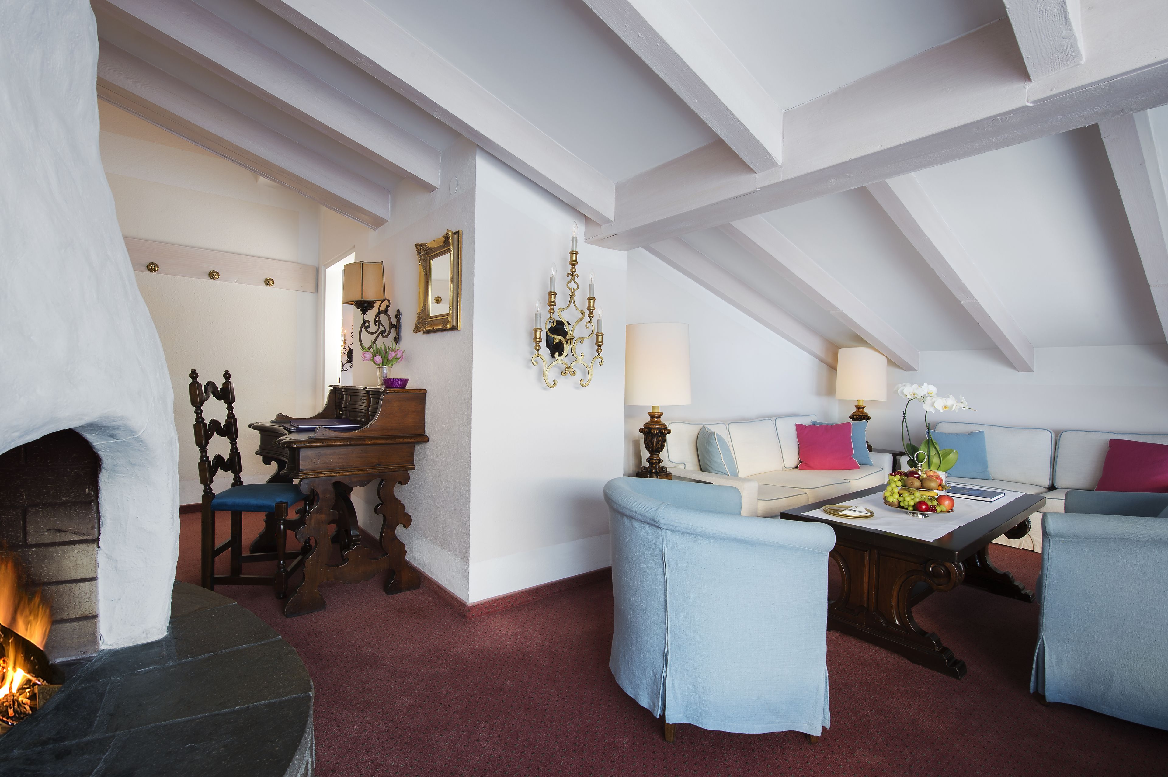 A cozy attic room with a fireplace, a piano, a sofa, and a dining table with fruit.