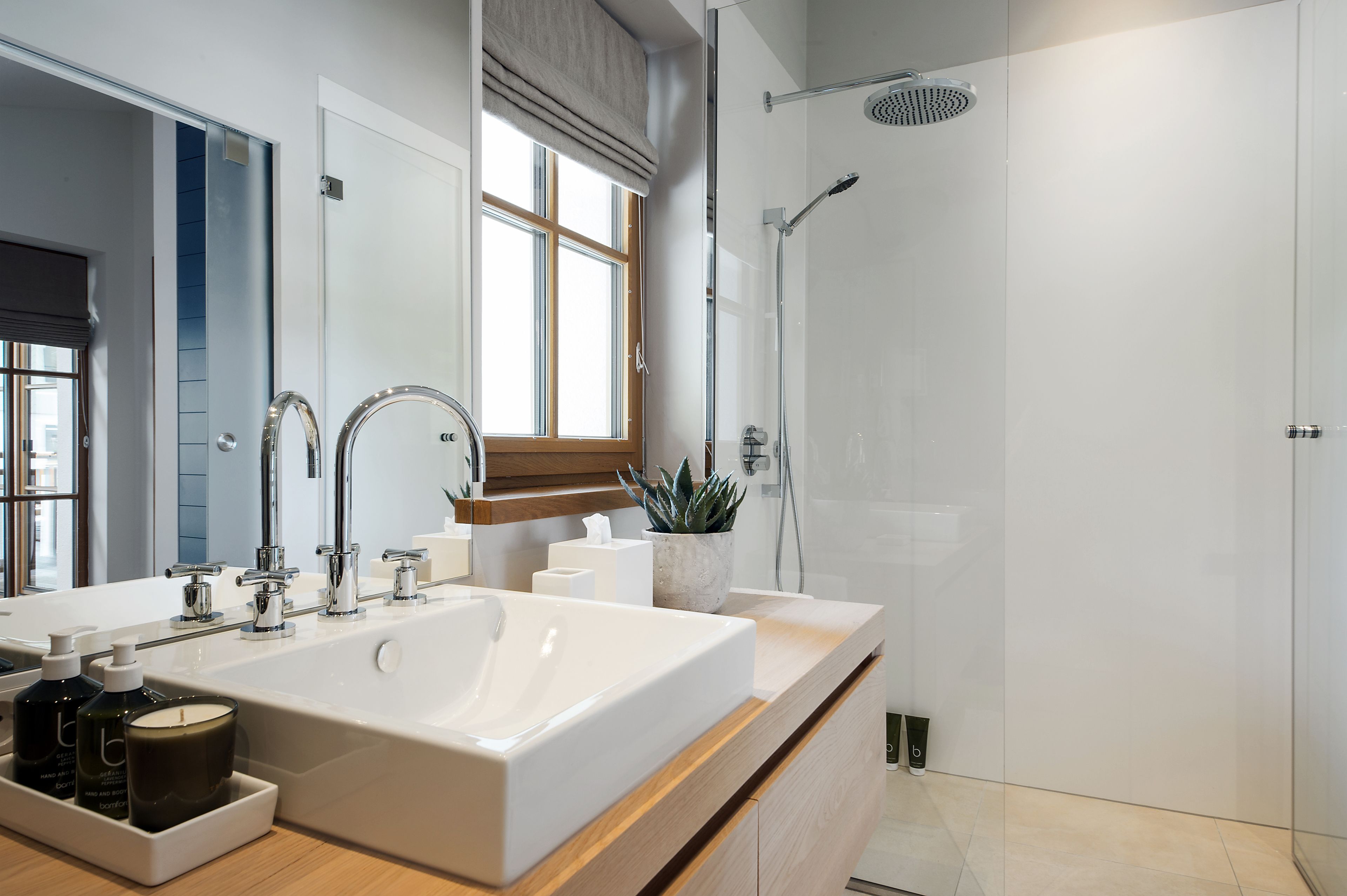 A modern bathroom with a large white sink, a mirror, a shower, and a window.