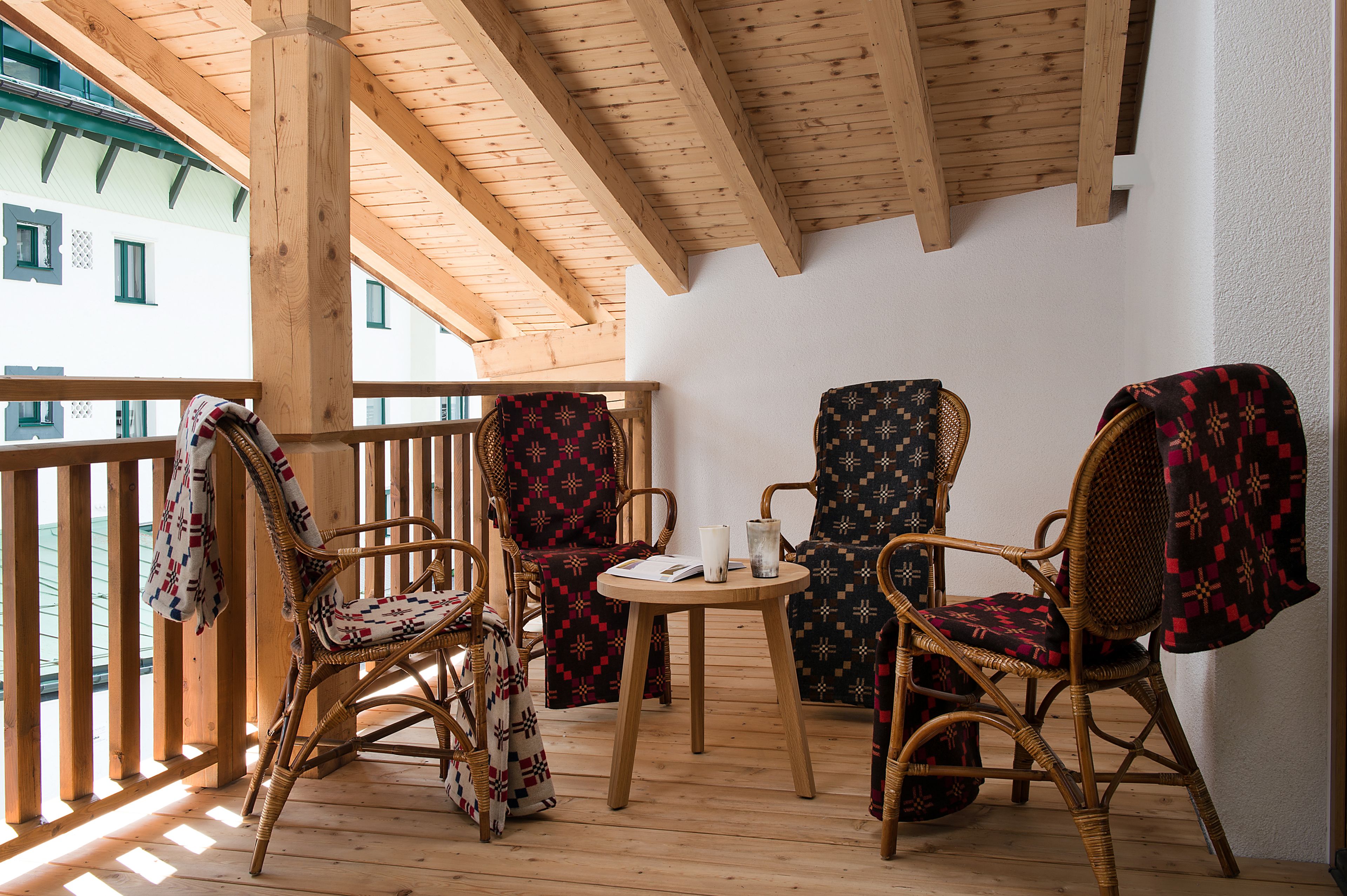 Una terraza de madera con sillas de mimbre y una pequeña mesa, con vista a un edificio con ventanas.