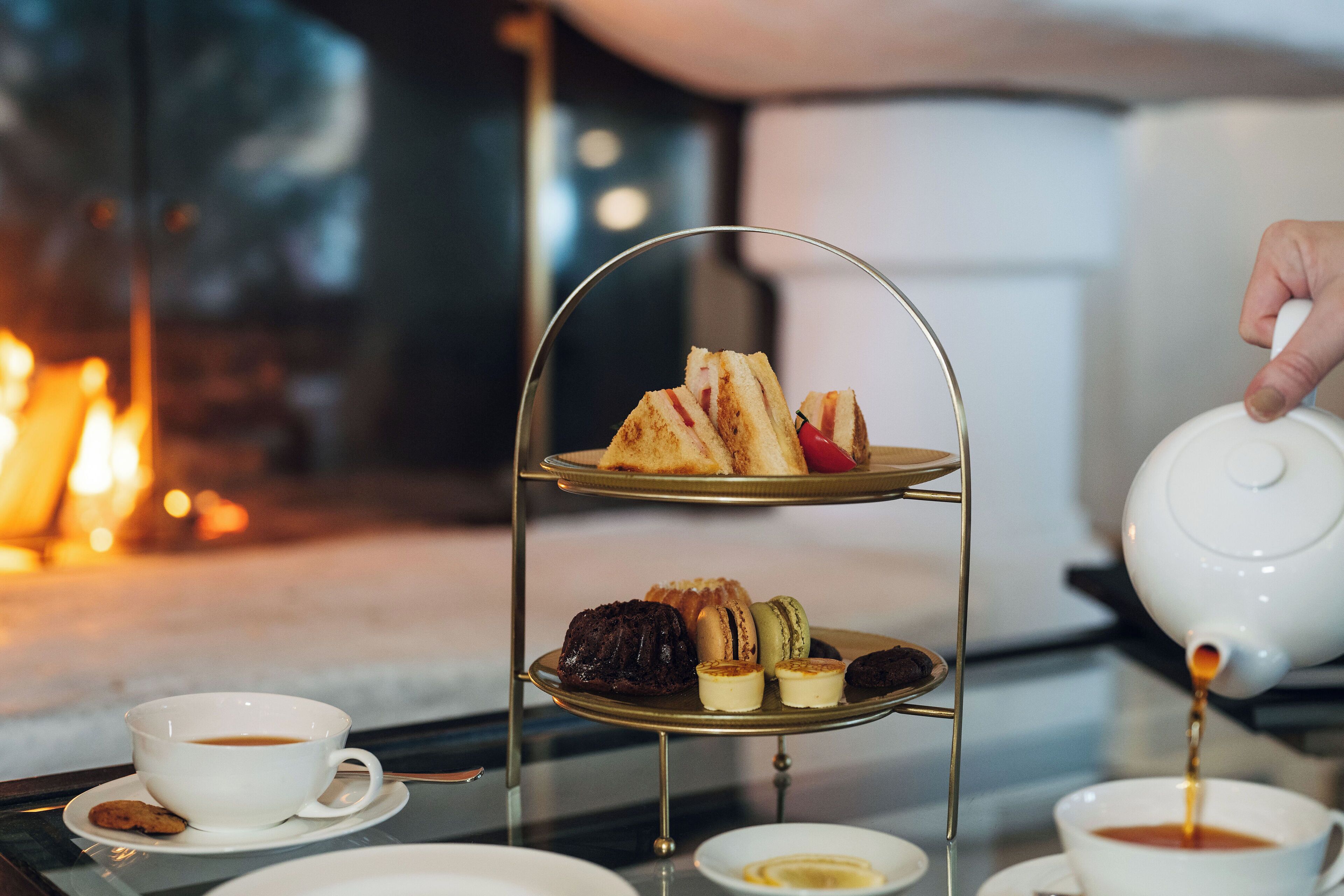 A three-tiered tea service with pastries and sandwiches, a cup of coffee, and a teapot pouring tea.