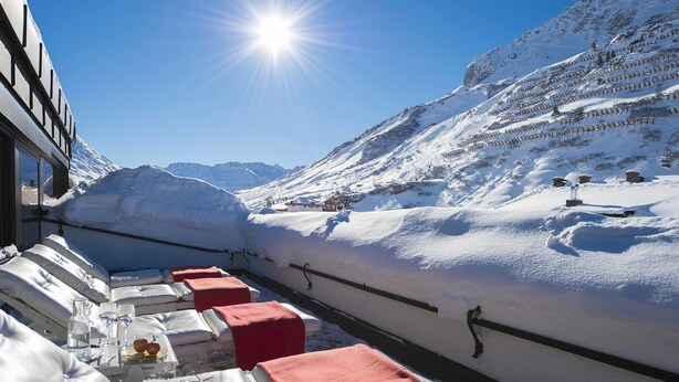 Un paisaje montañoso cubierto de nieve con un área de comedor que presenta mesas con cojines rojos y manteles blancos, vasos y una botella.