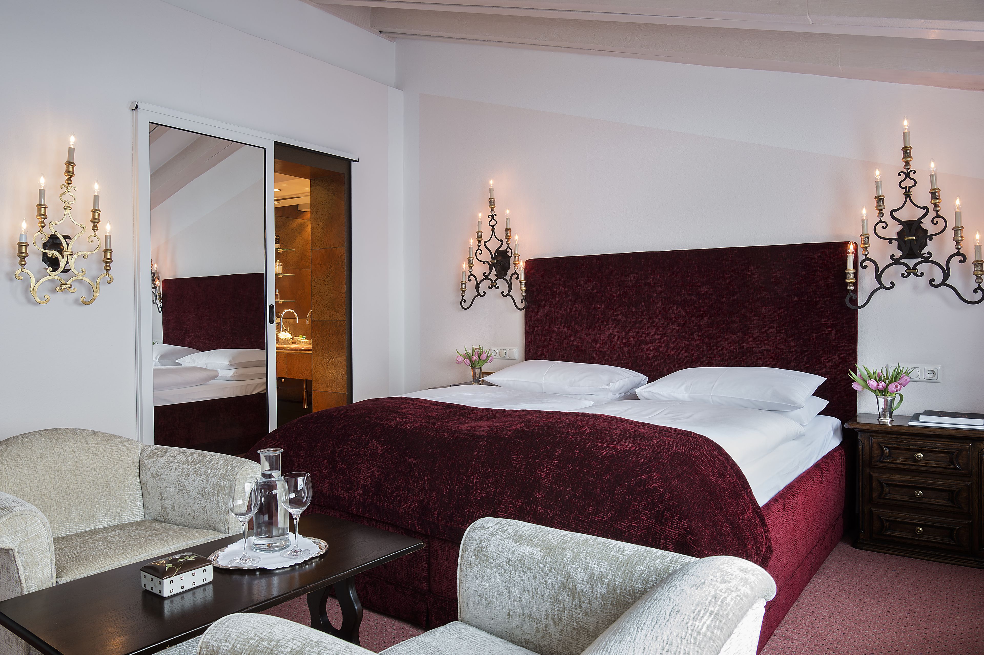 A hotel room with a large bed, a sofa, a table with glasses and a bottle, and two wall sconces.