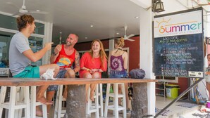 Um grupo de pessoas sentadas em um bar com uma lousa listando comodidades como dormitórios de 6 a 8 camas, ar condicionado e Wi-Fi gratuito.