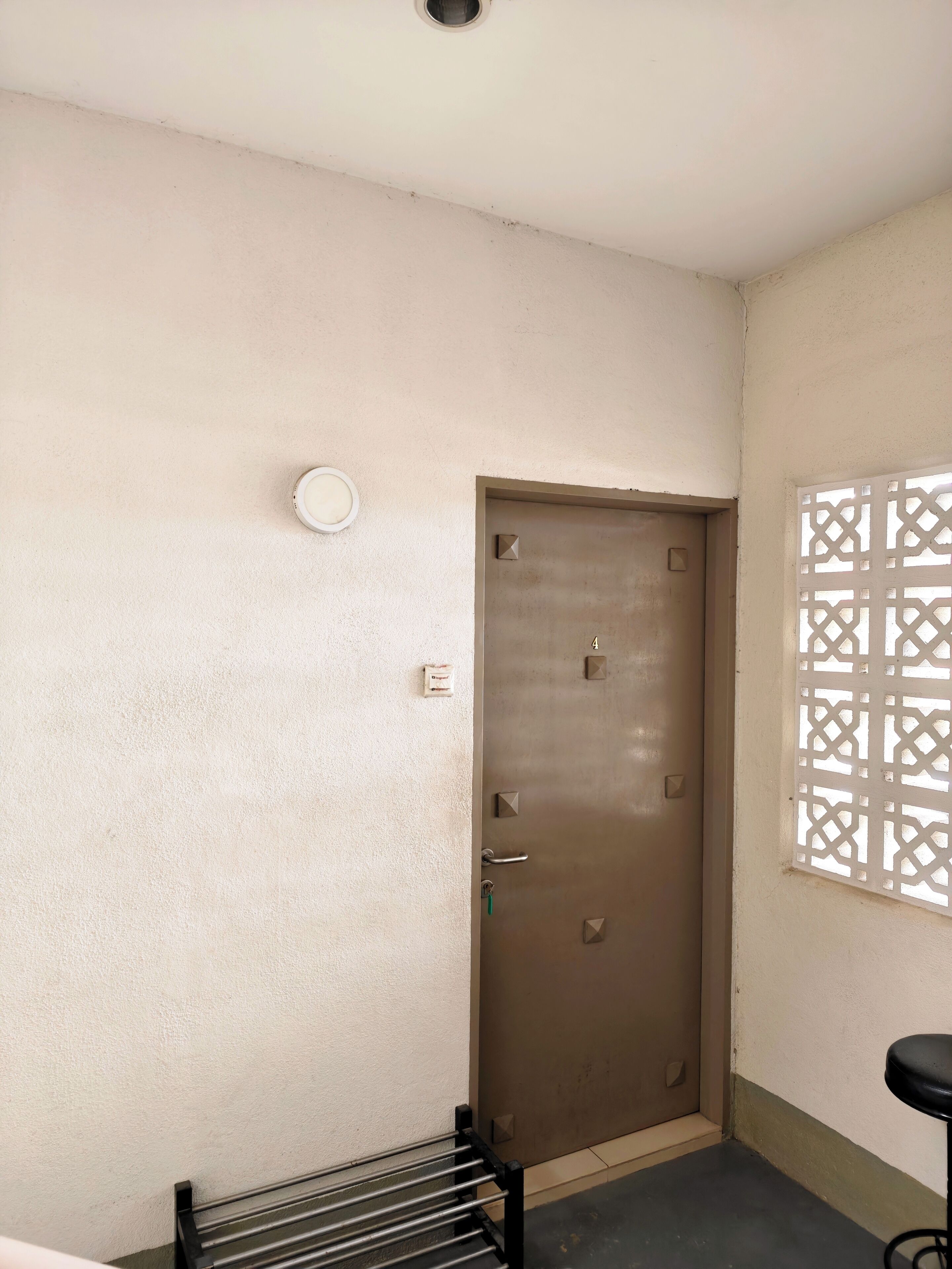 Una habitación con una puerta marrón, una ventana pequeña con un diseño de celosía y un estante de metal negro.