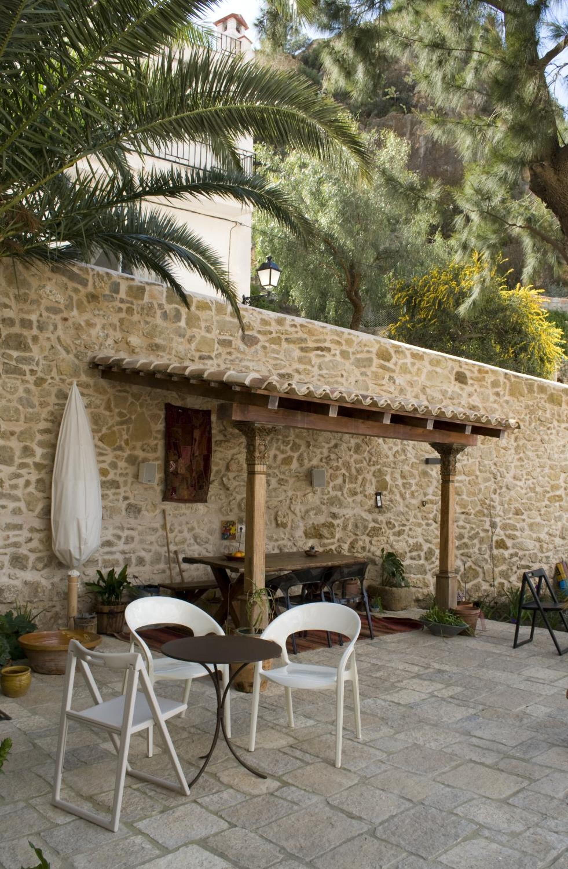 A stone patio with two white chairs and a table, surrounded by potted plants and a pergola.