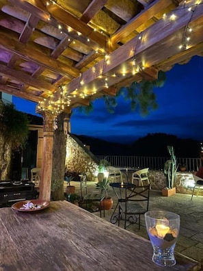 Un espacio de comedor al aire libre con una pérgola de madera, luces colgantes y plantas en macetas.