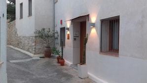 A narrow, paved courtyard with a white building featuring a wooden door and windows. There are potted plants and a stone wall visible. - La Baranda (Chulilla)