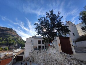 Un edificio de piedra con un árbol grande en primer plano, cielo despejado y montañas al fondo.