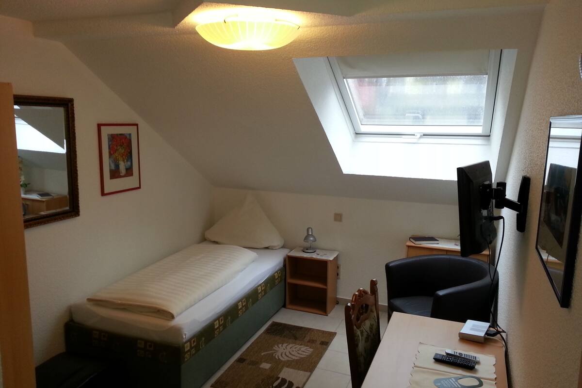 A small, well-lit room with a single bed, a desk with a computer, a chair, and a TV.