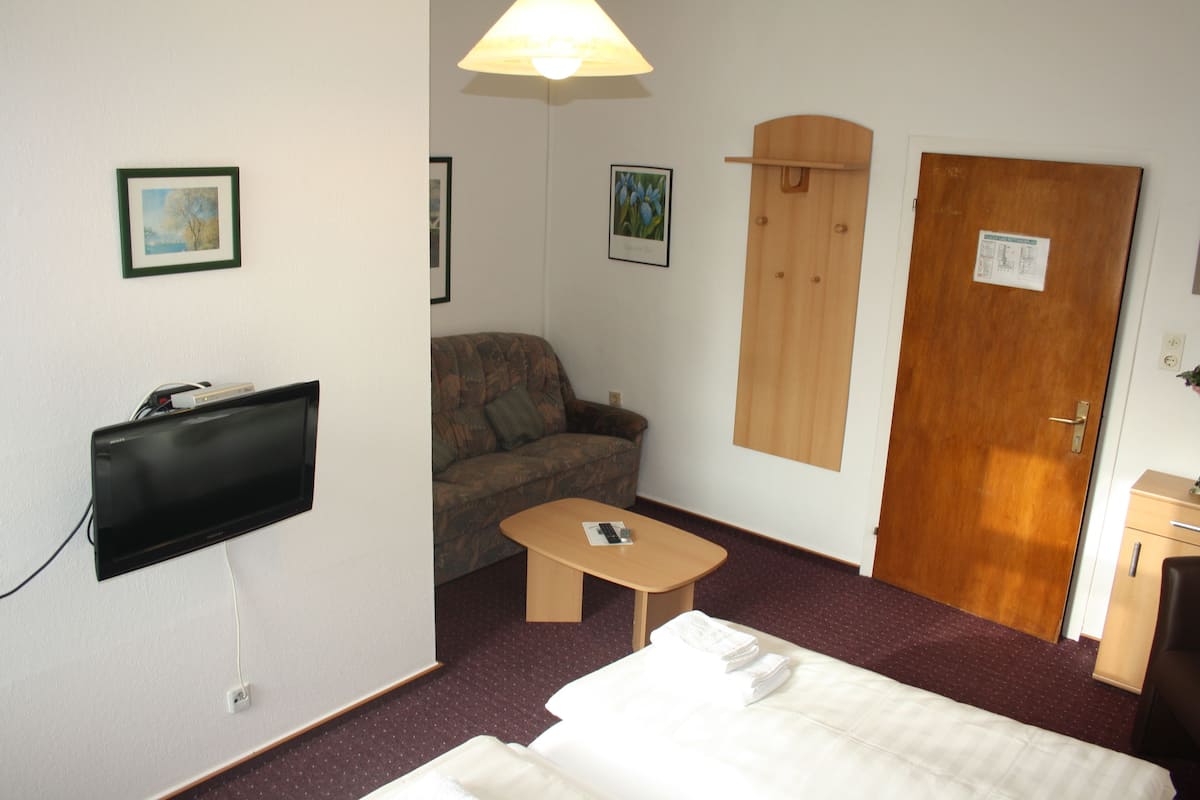 A hotel room with a bed, a sofa, a small table, a TV mounted on the wall, and a wooden door.