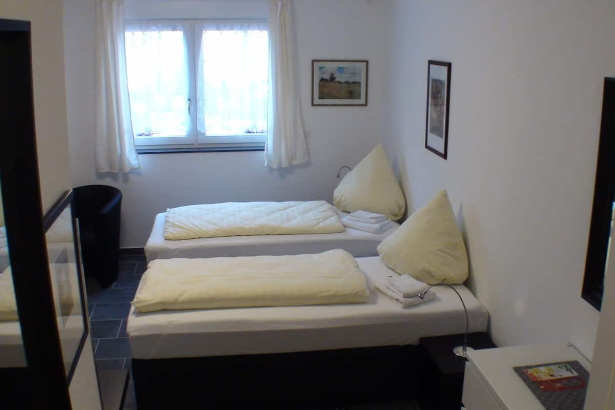 A small, clean room with two single beds, a window with curtains, a wall-mounted picture, and a trash bin.
