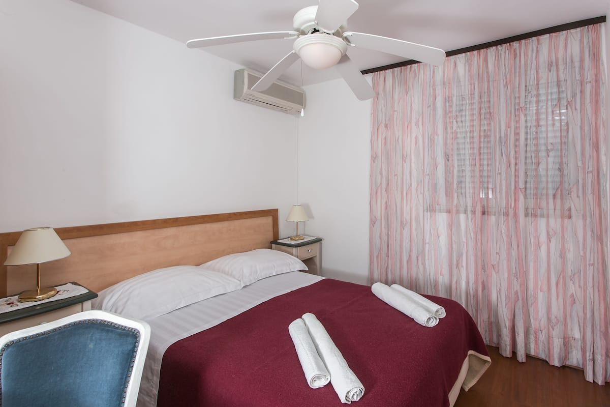 A hotel room with a bed, a ceiling fan, an air conditioning unit, a nightstand with a lamp, a chair, and a window with curtains.