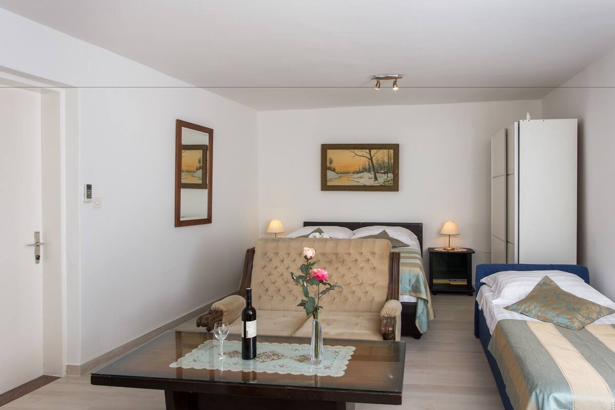 A hotel room with a bed, a sofa, a table with a bottle and a vase, a painting on the wall, and a mirror.