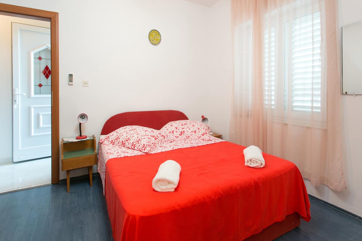 A hotel room with a red bed, white floral bedding, a wooden nightstand, a bedside table with a lamp, and a window with sheer curtains.