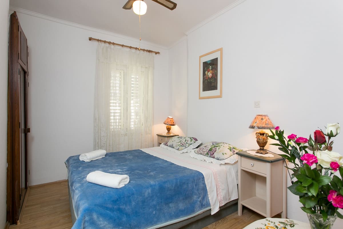 A bedroom with a bed, two bedside tables, a nightstand, a lamp, a vase with flowers, and a painting on the wall.