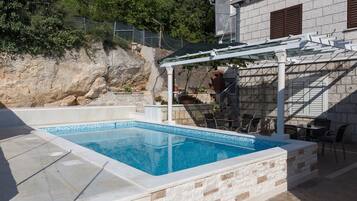 Een rechthoekig zwembad met een helderblauw binnengedeelte, omgeven door stenen muren en een pergola met een wit frame.