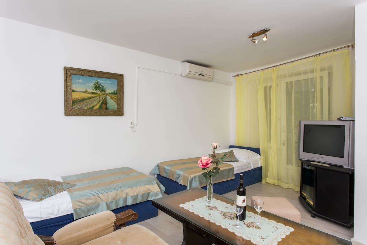 A room with two single beds, a sofa, a small table with a wine glass and bottle, a TV on a stand, and a painting on the wall.